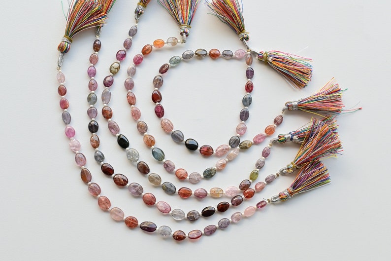 May include: A set of six strands of oval-shaped gemstone beads in various shades of pink, purple, blue, and green. Each strand has a colorful tassel at the end.