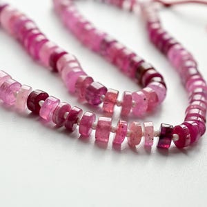 May include: A necklace composed of small, faceted beads in varying shades of pink and magenta. The beads are strung together, creating a flowing, organic shape. The background is a clean, white surface, highlighting the necklace's colors and form.