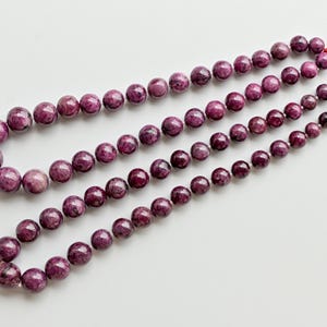 May include: Three strands of round, deep magenta gemstone beads. The beads vary in size, with the largest at the top and the smallest at the bottom. Each strand is strung on a red cord with a tassel at the end. The background is white.