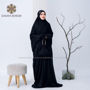 Może przedstawiać: Czarny str&oacute;j do modlitwy z pasującą torbą ze sznurkiem. Długa szata zakrywa głowę i ciało. Widoczna jest nazwa marki "KANAYA BORDIR". Mały, jasno zabarwiony stołek znajduje się po lewej stronie. Str&oacute;j jest noszony przez osobę.