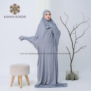 Może przedstawiać: Jasnoszary str&oacute;j modlitewny z pasującą osłoną głowy. Str&oacute;j jest długi i powł&oacute;czysty, z szerokimi rękawami. Nazwa marki "KANAYA BORDIR" jest nadrukowana na ubraniu. Mały, okrągły, beżowy stołek z drewnianymi nogami znajduje się w tle.