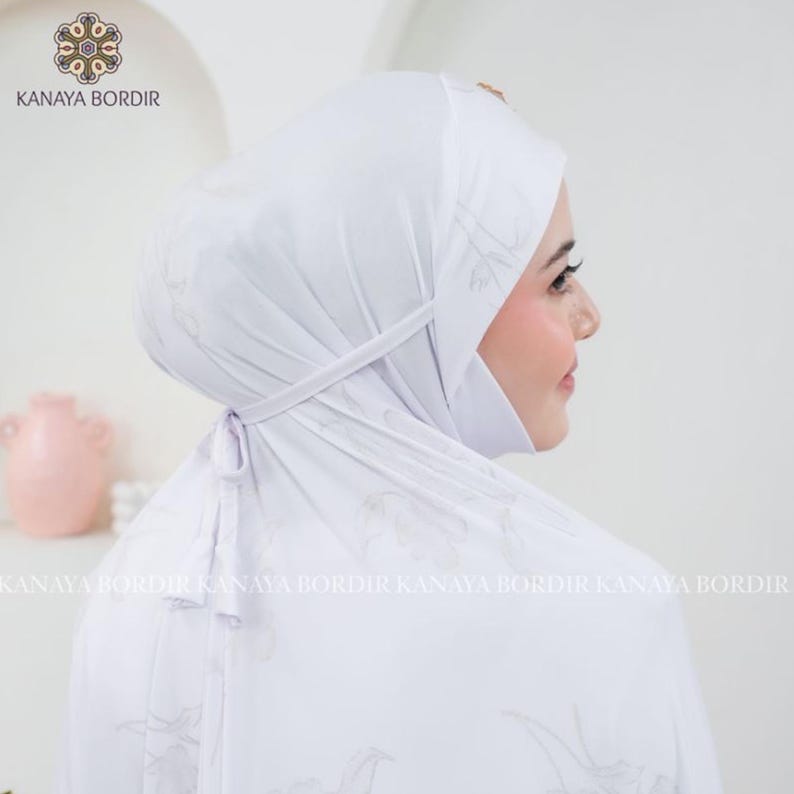 Może przedstawiać: Biała chusta na głowę z subtelnym kwiatowym wzorem i wiązaniem. Logo KANAYA BORDIR widoczne jest w lewym g&oacute;rnym rogu. Tkanina wydaje się miękka i dobrze się układa. Chusta przeznaczona jest do noszenia.