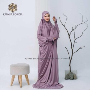 Może przedstawiać: Zestaw do modlitwy w kolorze malwy, składający się z długiej sukni i nakrycia głowy, z pasującą torbą ze sznurkiem. Widoczna jest nazwa marki "KANAYA BORDIR". W tle znajduje się mały, jasny stołek. Skromny styl.