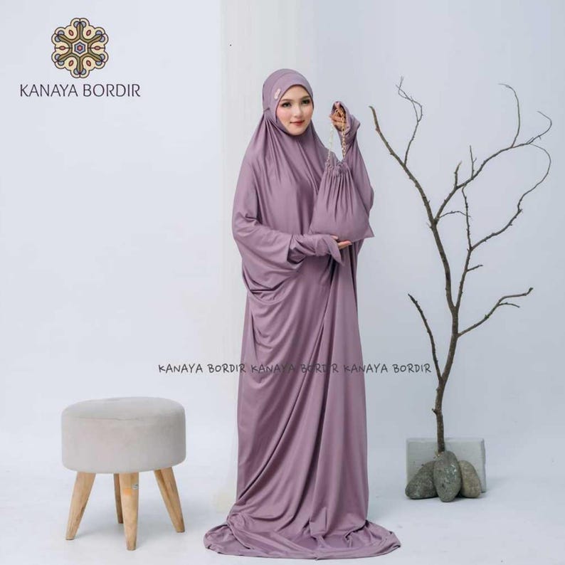 Może przedstawiać: Zestaw do modlitwy w kolorze malwy, składający się z długiej sukni i nakrycia głowy, z pasującą torbą ze sznurkiem. Widoczna jest nazwa marki "KANAYA BORDIR". W tle znajduje się mały, jasny stołek. Skromny styl.