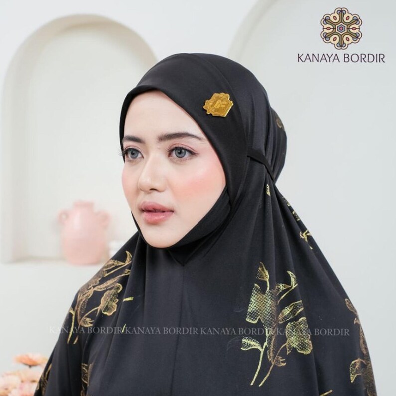 Może przedstawiać: Czarny hidżab ze złotym kwiatowym wzorem i złotą broszką. Hidżab jest noszony przez osobę. Tekst "KANAYA BORDIR" jest widoczny w prawym g&oacute;rnym rogu i na dole obrazu.