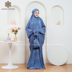 Może przedstawiać: Niebieski str&oacute;j modlitewny z pasującym nakryciem głowy i małą torbą ze sznurkiem. Tkanina ma subtelny kwiatowy wz&oacute;r w jaśniejszym odcieniu niebieskiego. Str&oacute;j jest noszony przez osobę stojącą przed białą ścianą.