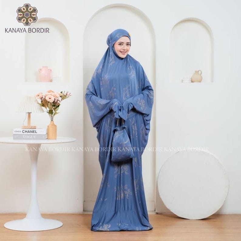 Może przedstawiać: Niebieski str&oacute;j modlitewny z pasującym nakryciem głowy i małą torbą ze sznurkiem. Tkanina ma subtelny kwiatowy wz&oacute;r w jaśniejszym odcieniu niebieskiego. Str&oacute;j jest noszony przez osobę stojącą przed białą ścianą.