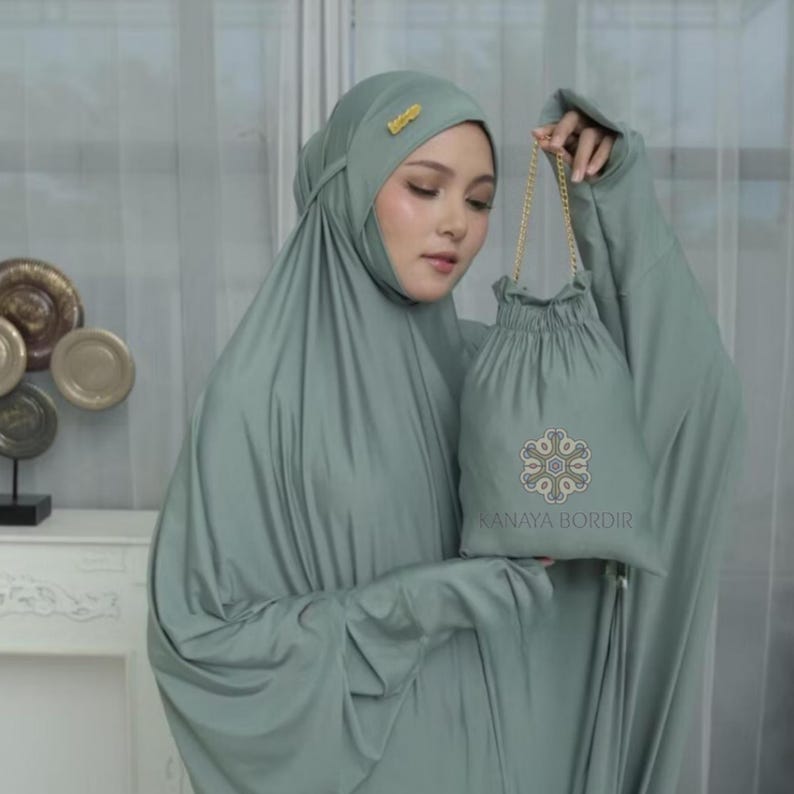 Może przedstawiać: Zestaw do modlitwy w kolorze szałwii, w tym nakrycie głowy i pasująca torba ze sznurkiem. Torba posiada haftowany wz&oacute;r i napis "KANAYA BORDIR". Zestaw wykonany jest z miękkiej, zwiewnej tkaniny.