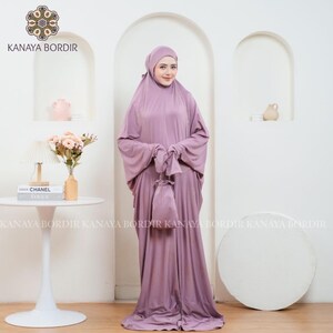 Może przedstawiać: Liliowy zestaw do modlitwy, w tym nakrycie głowy, sukienka z długim rękawem i pasująca torba ze sznurkiem. Zestaw wykonany jest z miękkiej, zwiewnej tkaniny. Marka "KANAYA BORDIR" jest widoczna u g&oacute;ry.