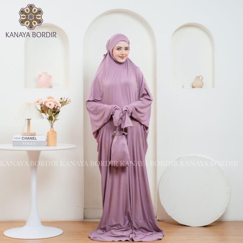 Może przedstawiać: Liliowy zestaw do modlitwy, w tym nakrycie głowy, sukienka z długim rękawem i pasująca torba ze sznurkiem. Zestaw wykonany jest z miękkiej, zwiewnej tkaniny. Marka "KANAYA BORDIR" jest widoczna u g&oacute;ry.