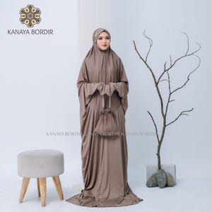 Może przedstawiać: Skromny brązowy str&oacute;j modlitewny z pasującą osłoną głowy i małą torbą ze sznurkiem. Str&oacute;j jest do ziemi i ma długie rękawy. Nazwa marki "KANAYA BORDIR" jest widoczna w lewym g&oacute;rnym rogu i na dole obrazu.