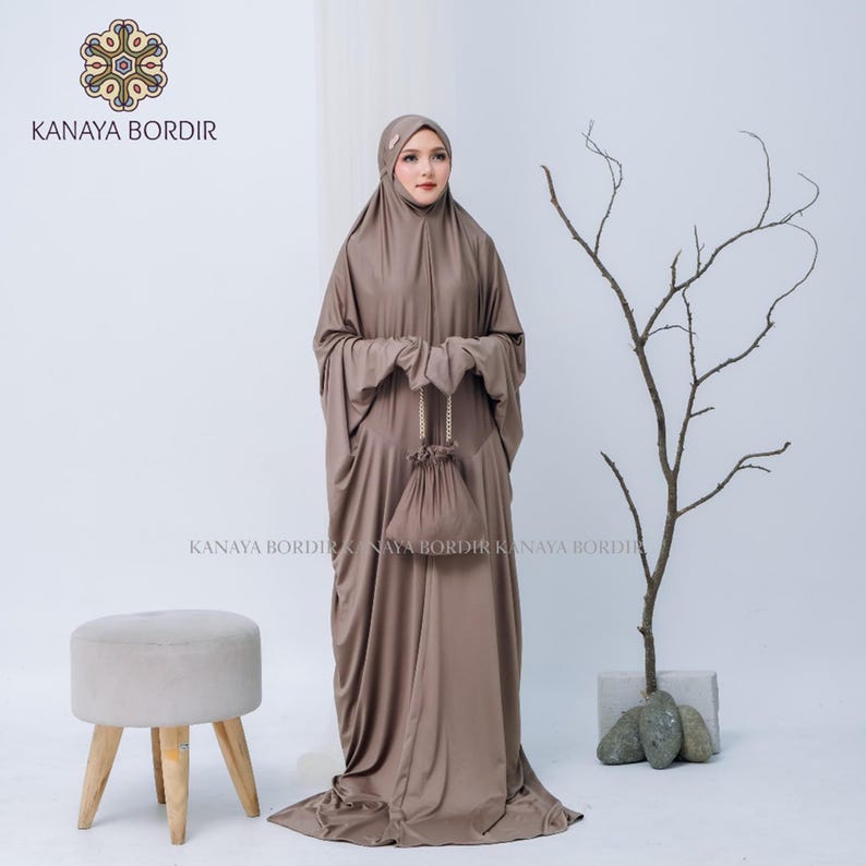 Może przedstawiać: Skromny brązowy str&oacute;j modlitewny z pasującą osłoną głowy i małą torbą ze sznurkiem. Str&oacute;j jest do ziemi i ma długie rękawy. Nazwa marki "KANAYA BORDIR" jest widoczna w lewym g&oacute;rnym rogu i na dole obrazu.