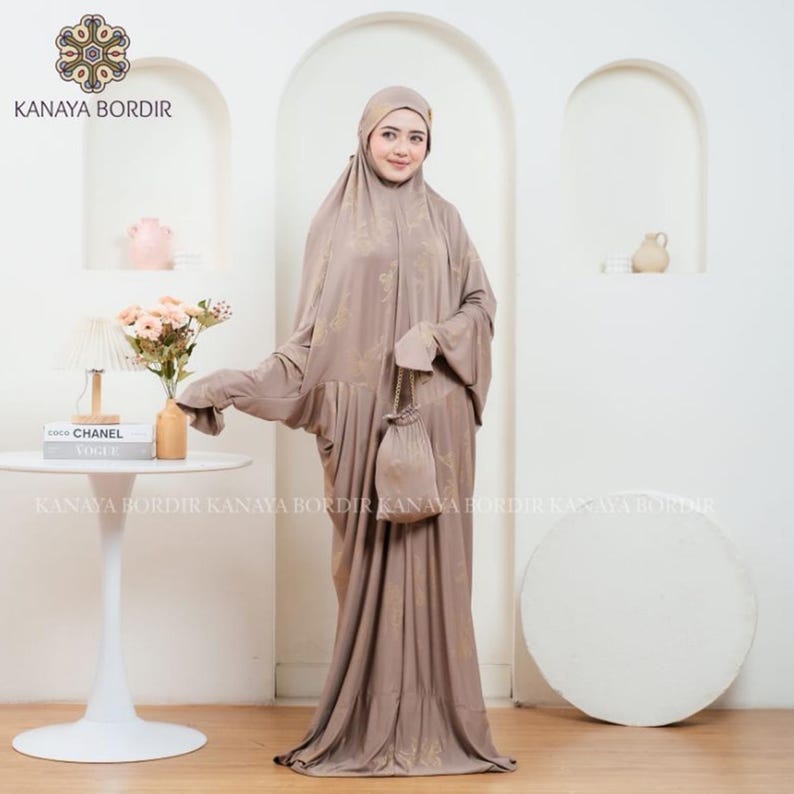 Może przedstawiać: Skromna sukienka w kolorze taupe z pasującą osłoną głowy i małą torebką. Sukienka ma subtelny kwiatowy wz&oacute;r i długie, zwiewne rękawy. Widoczna jest nazwa marki "KANAYA BORDIR".