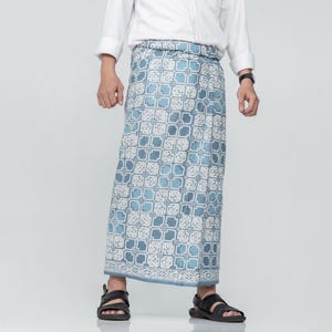 May include: A long, patterned sarong in shades of blue and white. The fabric features a repeating geometric design. The sarong is worn with a white button-down shirt and black sandals. The overall style is casual and comfortable.