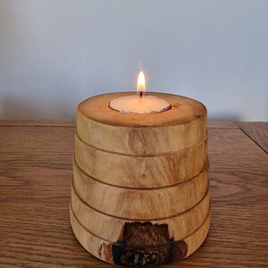 May include: A wooden candle holder with a natural wood finish. The holder has a cylindrical shape with a slightly flared top and a smooth, polished surface. A small white tea light candle is placed in the centre of the holder, and the candle is lit, casting a warm glow.