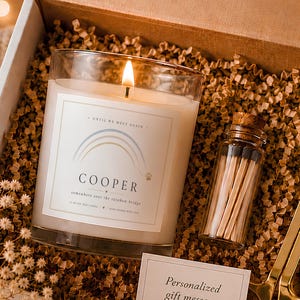 May include: A gift box containing a lit candle with a rainbow label reading Cooper, a small glass bottle of matches, and gold-toned metal wick trimmers. A small white card with the text Personalized gift message available is included.
