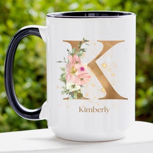 May include: White ceramic mug with a black handle. The mug features a gold letter K with pink and white floral accents and gold glitter. The name Kimberly is written below the letter.