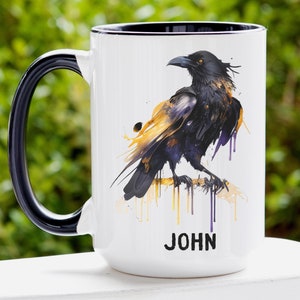 May include: White ceramic mug with a black handle and rim. The mug features a watercolor illustration of a black raven with yellow and purple accents. The name "JOHN" is printed in black at the bottom of the mug.
