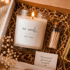 May include: A gift box containing a lit candle with a label reading no words... just hugs, a small glass bottle of matches, and gold-toned metal wick trimmers. A small card reads Personalized gift message available, all nestled in brown paper filler.