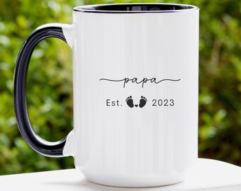 May include: White ceramic mug with a black handle and rim. The mug has black text that reads "papa est. 2023" with a small heart and two footprints between the words "est." and "2023".
