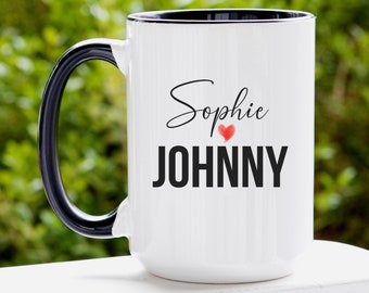 May include: White ceramic mug with a black handle and rim. The mug features the names "Sophie" and "JOHNNY" in black text, with a red heart between the names. The mug is designed for beverages.
