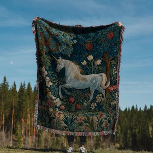 May include: A tapestry throw blanket featuring a white unicorn in a forest scene with flowers and trees. The blanket has a dark blue background with a fringed edge. The unicorn has a long horn and is in a running pose.
