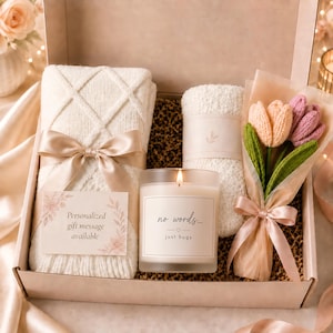 May include: A gift box containing a cream-colored knit blanket tied with a satin ribbon, a pair of fuzzy socks, a glass candle with a label reading no words... just hugs, and a bouquet of three crocheted tulips wrapped in translucent paper.