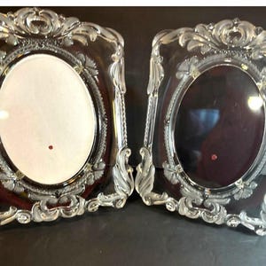 May include: Two rectangular picture frames with ornate, clear glass borders featuring sculpted floral and scrollwork details. Each frame has a central oval opening, one with a white insert and one with a dark maroon insert, both featuring a small red dot.