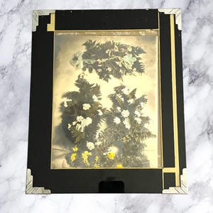 Può includere: Una fotografia antica incorniciata con una composizione floreale. La fotografia è racchiusa in una cornice nera con dettagli dorati e protezioni angolari argentate. La composizione floreale comprende fiori bianchi e gialli, con fogliame verde.