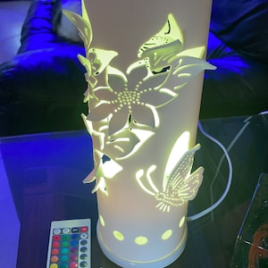 Può includere: Una lampada in ceramica bianca con un design floreale e farfalla. La lampada ha un telecomando con più luci colorate.