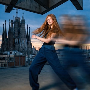 Può includere: Una persona con lunghi capelli rossi indossa un top nero e pantaloni blu, in piedi su un tetto con un effetto di movimento sfocato. Lo sfondo presenta un grande edificio con più guglie e un cielo nuvoloso.