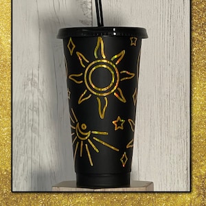 May include: Black reusable tumbler with a black lid and straw. The tumbler is decorated with a gold sun and star design. The tumbler is approximately 18 cm tall.
