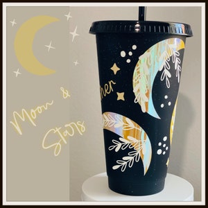 May include: Black tumbler with a lid and straw, decorated with a celestial design. The design includes a crescent moon, stars, and the words "Moon & Stars". The tumbler has a glittery finish and a holographic moon design.