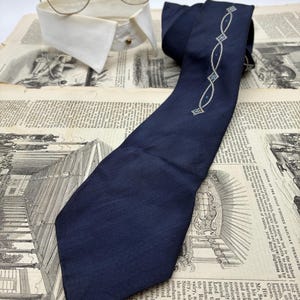 May include: A dark navy blue necktie featuring a thin, embroidered silver-toned chain pattern with small blue accents. The tie is positioned near a white detachable shirt collar and a pair of brown-rimmed eyeglasses with clear lenses.