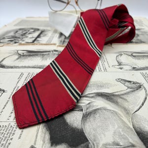 May include: A red necktie featuring diagonal stripes in black and cream, positioned alongside a pair of browline glasses with clear lenses. The necktie has a smooth texture and a pointed end, showcasing a classic pattern of repeating dark and light lines.