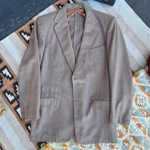 May include: A light brown blazer featuring a single-breasted front with a two-button closure, notched lapels, a chest pocket, two lower flap pockets, a small ticket pocket above the right flap pocket, and four buttons on each sleeve cuff.