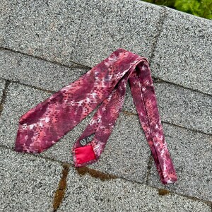 May include: A burgundy tie with a patterned design in shades of red and white. The tie is made of a silky material and has a red lining. The label reads "Cutler".