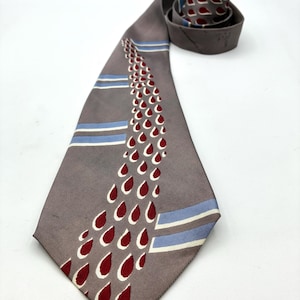 May include: A gray necktie with a repeating pattern of red teardrops outlined in white. The tie also features two horizontal stripes of blue and white. The tie is rolled up at the top.
