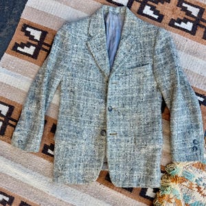 May include: A gray and white tweed blazer featuring a textured speckled pattern, a notched lapel, two front buttons, a single chest pocket, and two buttons on each sleeve cuff. The interior is lined with a solid gray fabric, providing a structured look.