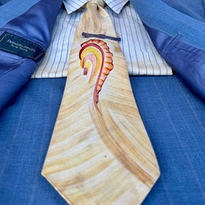 May include: A light blue suit jacket with a white striped shirt and a beige tie with a colorful abstract design. The tie has a silver tie clip. The label "Broadstreet's New York" is visible on the jacket.