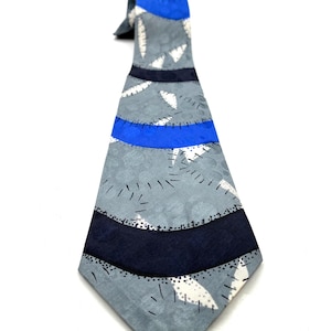 May include: A necktie featuring a gray base with a subtle circular pattern, accented by horizontal bands of bright blue and dark navy. The design includes white leaf-like shapes outlined with black dashed lines that mimic a stitched appearance.
