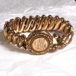 May include: A gold-toned metal bracelet featuring a flexible, expandable link band and a central circular clasp. The clasp is decorated with an ornate, embossed floral border and a smooth, round center engraved with the stylized cursive letters LR.