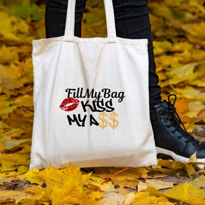 Könnte beinhalten: Cremefarbene Tragetasche mit schwarzem Text und roter Lippen-Grafik. Der Text lautet "Fill My Bag Kiss My A$$" mit goldenen Dollarzeichen. Die Tasche wird von einer Person in schwarzer Hose und schwarzen Stiefeln gehalten.