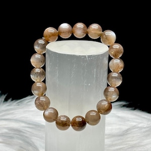 May include: A bracelet of light brown, spherical beads, each with unique colour and texture variations. The bracelet is presented on a white, cylindrical stand, set against a black backdrop and a fluffy white surface.