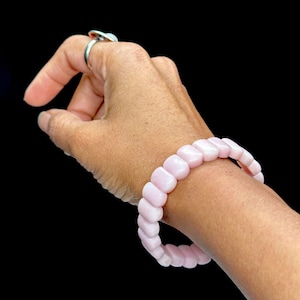 May include: A pink gemstone bracelet with square-shaped beads. The bracelet is made of natural stones and is worn on a person's wrist.