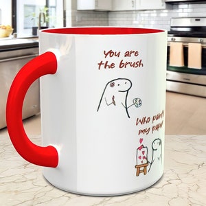 May include: White ceramic mug with a red handle and red rim. The mug has a black and white illustration of a person holding a paintbrush and a canvas. The text on the mug reads "You are the brush. Who paints my paper?"