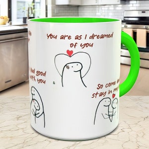 May include: White ceramic mug with a green rim and handle. The mug has a black and white illustration of two stick figures with hearts and the text "You are as I dreamed of you", "I feel good with you", and "So come and stay in my heart".