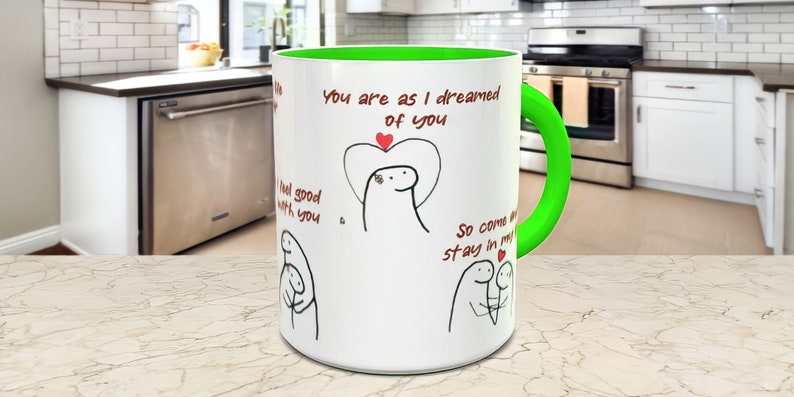 May include: White ceramic mug with a green rim and handle. The mug has a black and white illustration of two stick figures with hearts and the text "You are as I dreamed of you", "I feel good with you", and "So come and stay in my heart".