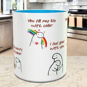 May include: White ceramic mug with a blue rim. The mug features a hand-drawn illustration of a person with a rainbow above their head and the text "You fill my life with color". There is also a hand-drawn illustration of two people hugging with the text "I feel good with you".