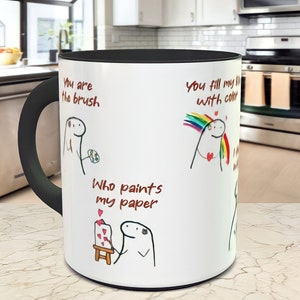 May include: White ceramic mug with a black handle and black interior. The mug has a cartoon illustration of a person with the text "You are the brush", "You fill my life with color", and "Who paints my paper".